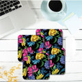 A set of four square floral pattern coasters with a glitter finish, displayed on a wooden surface alongside a cup of coffee and a laptop.